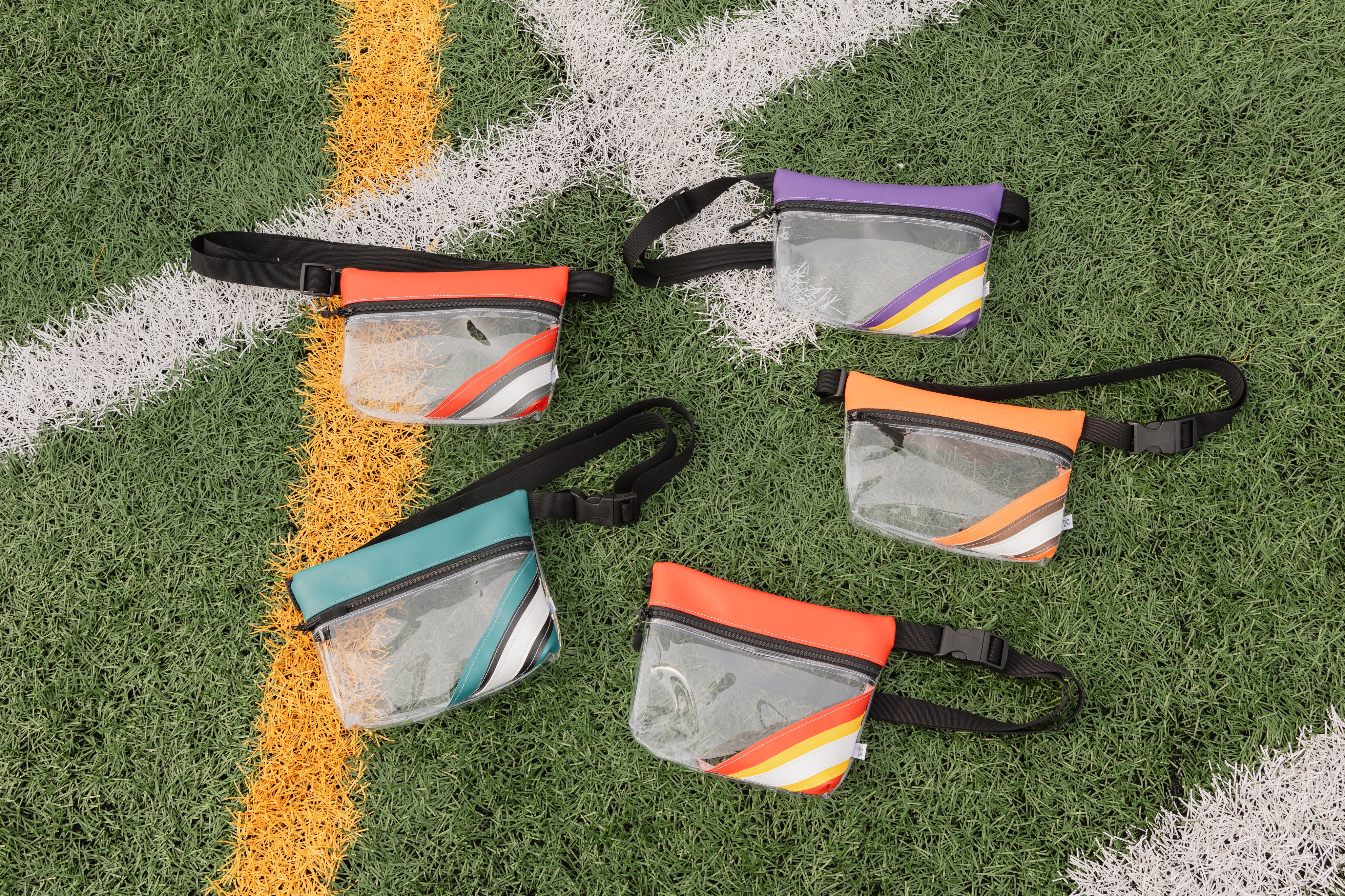 Five clear waterproof pouch fanny packs with colorful striped accents and black straps, arranged on a green turf field with white and yellow lines.